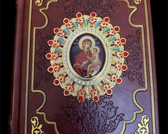 May include: A brown leather-bound book featuring an ornate gold-toned oval centerpiece with a central religious icon of a figure holding a child. The centerpiece is decorated with red and turquoise stones and clear crystals, surrounded by gold filigree.