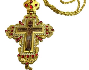 Może przedstawiać: Złoty metalowy krzyż pektoralny na pasującym łańcuszku typu lina. Krzyż posiada ozdobny filigranowy wzór, wysadzany małymi przezroczystymi cyrkoniami i prostokątnymi czerwonymi kamieniami. Centralna ikona przedstawia scenę ukrzyżowania Jezusa.