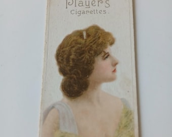 May include: A vintage Player's Cigarettes card featuring a woman with a light brown updo, wearing a white and yellow dress. The card reads "Player's Cigarettes" and "Gallery of Beauty No. 18."