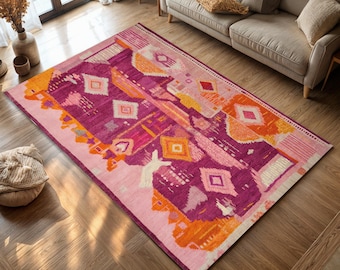 May include: A rectangular area rug with a vibrant geometric design. The rug features a palette of pink, purple, and orange hues, with diamond and abstract shapes. The rug is placed on a wooden floor, near a sofa.