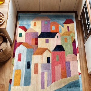 Peut inclure: Un tapis coloré avec un motif de ville avec des maisons dans différentes nuances de bleu, vert, rouge, jaune et violet. Le tapis a un aspect texturé et tissé.