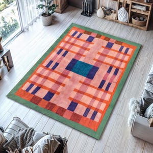 May include: A rectangular rug with a geometric design in pink, orange, blue, and green. The rug has a central teal square, a grid of orange and blue rectangles, and a green border. It is placed on a light wood floor, with a window in the background.