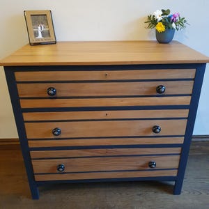 May include: Wooden chest of drawers with a light brown top and drawers. The drawers are light brown with dark blue accents and black knobs. A small framed photo and a vase of flowers sit on top.