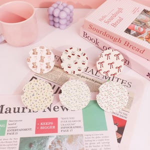 May include: A collection of six round car coasters with various patterns, including teddy bears, cherries, and bows. The coasters are on a light pink surface with a pink mug, a purple candle, and books in the background.