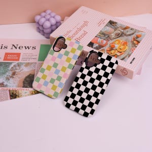 May include: Two phone cases with checkerboard patterns. One case has a black and white checkered design, and the other has a pastel checkered pattern in pink, yellow, blue, and green. A book with the text "Hello, Sourdough Bread" is in the background.