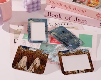 May include: Four rectangular badge holders with different designs. One features a cityscape, another has two ghosts in a forest, and two have a blank white space. The holders are on a newspaper with books in the background.