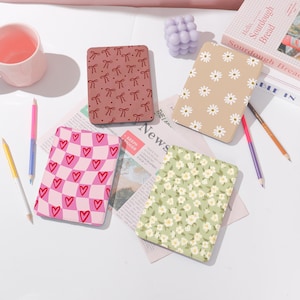May include: Four different patterned Kindle e-reader covers. One cover has a pink and white checkered pattern with red hearts. Another cover has a brown background with red bows. A third cover has a beige background with white daisies. The final cover has a green background with white flowers.