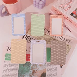 May include: Six ID badge holders in pastel shades of blue, green, peach, yellow, lavender, and brown. Each has a white interior and small white polka dots. They are arranged on a newspaper.