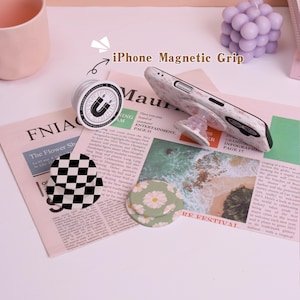 May include: An iPhone magnetic grip with a marble-patterned phone case, a black and white checkered grip, and a daisy-patterned grip. The text "iPhone Magnetic Grip" is visible. The items are on a newspaper.