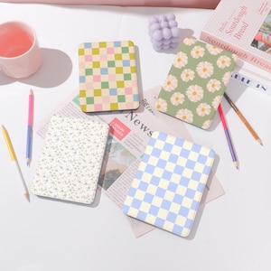 May include: Four different patterned e-reader cases. The cases are in a checkered pattern, a floral pattern, a checkered pattern, and a floral pattern. The cases are on a white surface with a newspaper, pencils, and a book.