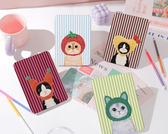 May include: Four rectangular cat-themed notebooks with striped backgrounds. Each notebook features a different cat wearing a hat: a tomato, a yellow bonnet, an orange hat, and a green cat hat. The notebooks are on a white surface.