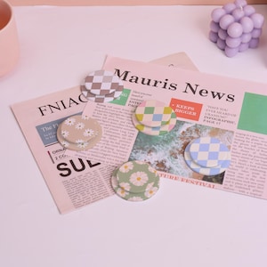 May include: Several round phone grips with various patterns, including daisies and checkered designs in different colours. The grips are displayed on a newspaper background. Measurements are not applicable.