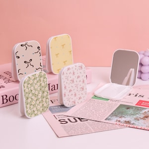 May include: Several small, rectangular compact mirrors with different patterns. The patterns include bows, floral designs, and a yellow background with bows. One mirror is open, showing a reflective surface.