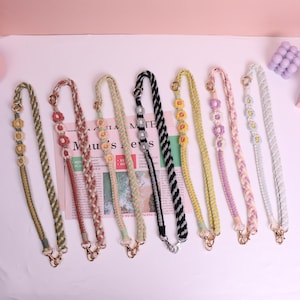 May include: A collection of eight colorful, braided phone straps with floral accents. Each strap features a different color scheme, including green, red, yellow, black, and pastel shades. The straps have gold or silver-toned clasps.