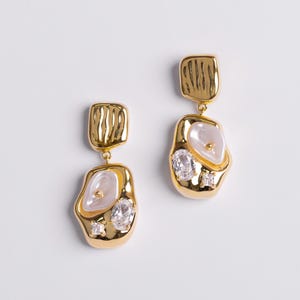 May include: A pair of gold-coloured drop earrings. Each earring has a rectangular top with a textured design, connected to a freeform pendant. The pendants have a pearl-like centre, clear crystals, and a small square crystal.