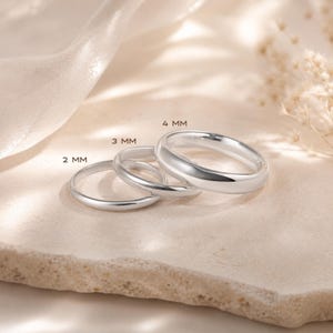 May include: Three polished silver-toned rings with smooth, rounded bands. The rings are arranged in a row, labelled with text indicating widths of 2 mm, 3 mm, and 4 mm. Each ring features a simple, solid design with a reflective metallic surface.