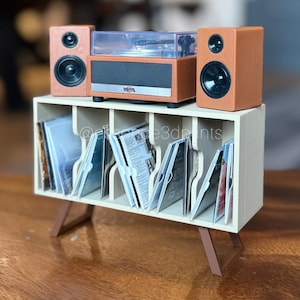 May include: A miniature record player with two brown speakers sits atop a cream-colored storage unit with five vertical slots holding small square discs. The record player has a clear plastic lid and a silver front panel with the word vinyl printed.