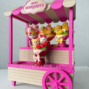 May include: A miniature pink and beige wooden cart with a striped awning and a sign reading MINI BOUQUETS. The cart displays several small bouquets featuring cartoon animal figurines, including bears and chicks, wrapped in decorative paper.