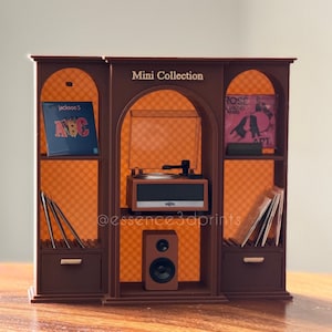 May include: A brown miniature shelving unit labeled Mini Collection featuring a central record player, a speaker, and side compartments. The shelves display miniature vinyl records including Jackson 5 and Rose, with additional records stored in slots.