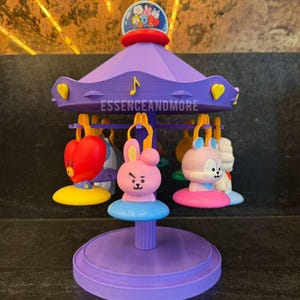 May include: A purple carousel toy featuring BT21 character figures hanging from yellow clips. The top canopy displays the text ESSENCEANDMORE and a small circular graphic. The carousel has a central pillar and a round base, with colorful character charms.