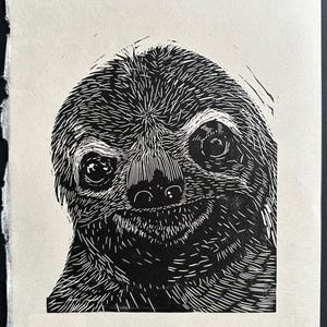 Pode incluir: Impressão em linóleo preto e branco do rosto de um bicho-preguiça. O bicho-preguiça tem um sorriso largo e cheio de dentes e seus olhos estão abertos e olhando diretamente para o espectador.