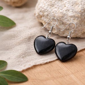 May include: A pair of black heart-shaped earrings with silver hooks. The earrings are displayed on a beige cloth next to a light-colored rock and green leaves, set against a wooden surface.