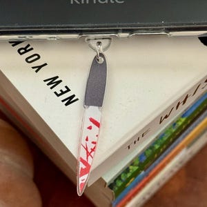 May include: A clear acrylic knife-shaped keychain with a gray handle and a red blood splatter design on the blade. The keychain is attached to a silver-colored ring. The word "kindle" is visible in the background.