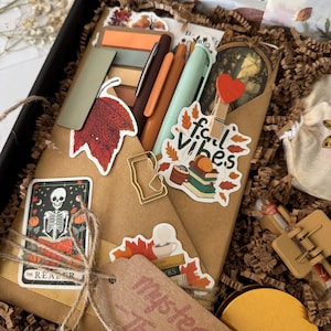 May include: A gift box with autumn-themed stationery and accessories. Inside, a brown envelope is decorated with stickers saying "fall vibes" and "mystery thriller". The box is tied with twine and a tag, creating a seasonal aesthetic.