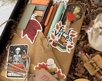 May include: A gift box filled with brown paper crinkle filler containing a wrapped book, a brown tag labeled Mystery Thriller, pens, sticky notes, a gold paperclip, and various autumn-themed stickers including a skeleton and the text Fall Vibes.