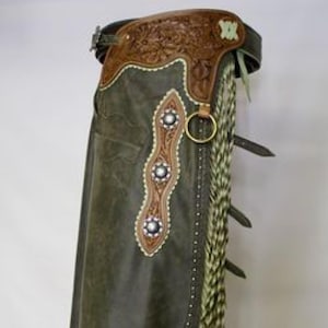 Pode incluir: Chaps de couro verde-oliva com detalhes em couro castanho e detalhes florais. Apresenta tachas decorativas prateadas, um anel dourado e franjas longas em tons de verde e creme. Uma peça de vestuário em estilo western.