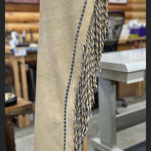 May include: A tan suede leather chap featuring a dark brown floral tooled leather yoke and lower accent panel. The edges are lined with small silver-toned metal studs, and a long fringe made of thin, braided leather strips runs down the side.