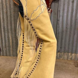 May include: Tan suede cowboy chaps with decorative stitching and fringe. The chaps have a light tan color with darker brown accents and a decorative pattern along the edges. The fringe hangs down the sides.