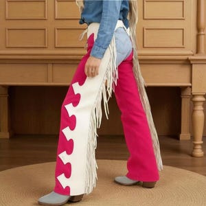 Puede incluir: Chaps de color rosa intenso con diseño de piezas de rompecabezas blancas y flecos. Los chaps se llevan sobre unos vaqueros azules. La persona lleva botas grises. El fondo es una pared de madera y una alfombra marrón.