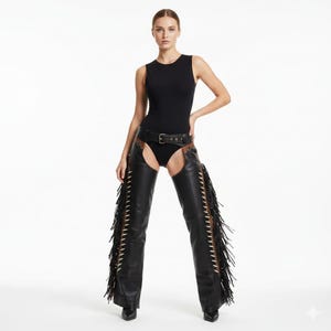 Puede incluir: Un body negro sin mangas combinado con unos chaps de cuero negro con flecos en los laterales. Los chaps se aseguran con un cinturón negro. La modelo posa sobre un fondo blanco.