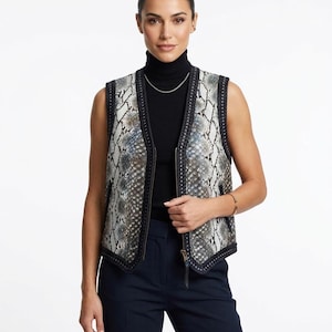 Puede incluir: Un chaleco sin mangas con estampado de piel de serpiente en tonos grises, blancos y azules. El chaleco tiene un ribete negro y cierre de cremallera. La modelo lleva un cuello alto negro y pantalones azul marino.