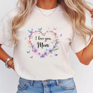 May include: White t-shirt featuring a floral heart design with the text "I love you, Mom." The heart is composed of pink and purple flowers, green leaves, and butterflies. The shirt is a casual, everyday wear item.