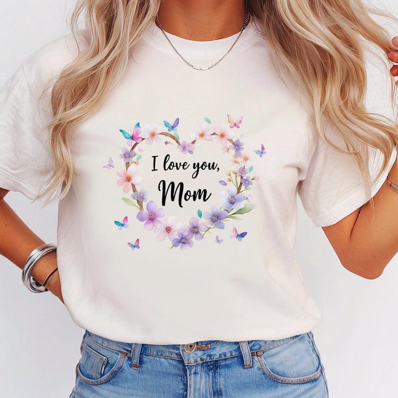 May include: White t-shirt featuring a floral heart design with the text "I love you, Mom." The heart is composed of pink and purple flowers, green leaves, and butterflies. The shirt is a casual, everyday wear item.
