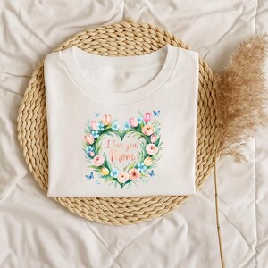 May include: White t-shirt with a floral heart design. The heart is composed of pink, yellow, and blue flowers, with the words "I love you, Mom" in the center. The shirt is on a woven mat.