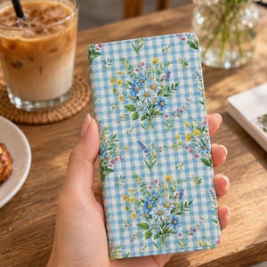 May include: A rectangular phone case featuring a light blue and white gingham check pattern overlaid with a repeating print of small wildflower bouquets in shades of blue, yellow, and green. The case has a smooth, flat surface and rounded edges.
