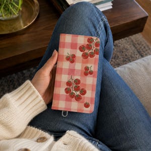 May include: A rectangular wallet featuring a light pink and white gingham check pattern decorated with clusters of small red tomatoes on green stems. The wallet has a smooth, flat surface and a small metal loop attached to the bottom edge.