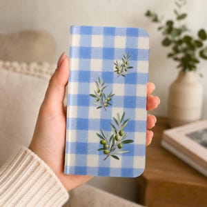 May include: A rectangular phone case featuring a light blue and white gingham check pattern. The design is decorated with three vertical illustrations of green olive branches with small green olives, arranged in a column down the centre.