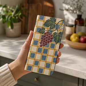 May include: A rectangular phone case featuring a blue and cream checkered pattern with a cluster of purple grapes and green leaves printed on the front. The design has a textured appearance, and the case is held in a vertical orientation.