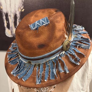 Puede incluir: Un sombrero fedora de fieltro marrón con acabado desgastado, adornado con una banda de mezclilla azul deshilachada y flecos. Un parche rectangular de mezclilla está cosido en la copa y una pluma oscura se inserta tras un emblema metálico color bronce.