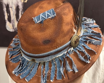 May include: A brown felt fedora hat featuring a distressed finish, adorned with a frayed blue denim band and fringe trim. A rectangular denim patch is stitched to the crown, and a single dark feather is tucked behind a circular bronze-toned metal emblem.
