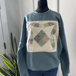 May include: A slate blue crewneck sweatshirt featuring a square patchwork quilt block on the chest. The quilt block is composed of various floral and plaid fabric squares in shades of green, cream, and blue, bordered by cream-colored lace trim.