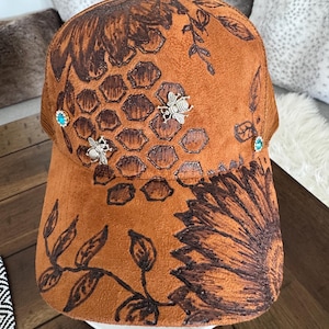 Op de afbeelding: Een bruine baseballpet met een donkerbruin, met de hand getekend zonnebloem- en honingraatpatroon. De pet is versierd met twee zilverkleurige bijenbedels en twee kleine ronde studs met blauwe en groene strass-steentjes op de voorpanelen.