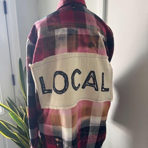 May include: A long-sleeve button-down flannel shirt featuring a red, black, and white plaid pattern with a bleached effect. A rectangular, off-white fabric patch is stitched onto the back with the word LOCAL printed in bold black letters.