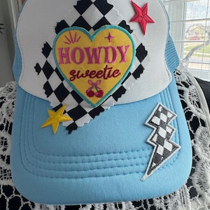 Peut inclure: Casquette de camionneur bleu clair avec un dos en filet blanc et une visière incurvée. Le chapeau présente un écusson en forme de cœur avec les mots "HOWDY sweetie" et des graphiques de cerises. Écussons supplémentaires : étoile rouge, étoile jaune et éclair à carreaux.