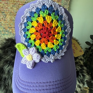 May include: A purple baseball cap with a vibrant, crocheted floral design. The flower features a rainbow of colors, including red, orange, yellow, green, and blue. A small crocheted lavender flower and green leaf accent the cap's side.