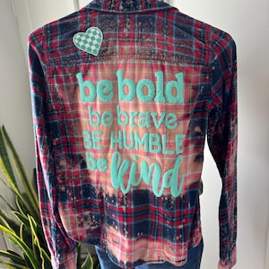 May include: A long-sleeve button-down flannel shirt with a red, blue, and white plaid pattern featuring a bleached effect. The back displays a teal checkered heart patch and the teal text: be bold, be brave, BE HUMBLE, be kind.